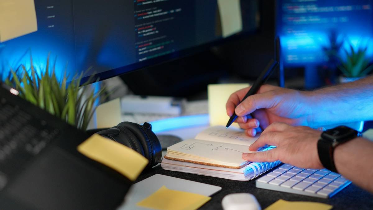 Check Programmer Project Notes Task Planning Workspace hand marking checkboxes in notebook with code screens and modern desk setup. Concept of software development organization, productivity and agile workflow